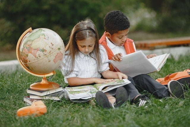 Niño y niña estudiando sobre el cesped durante su vuelta al cole mientras a su lado hay una pila de material escolar.